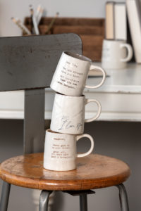 Dear You Mug - Strength. A stack of three cream ceramic textured coffee mugs, each with different color inner lining and message. One with a message about daughter, one engraved with a message about "you", and one reading "PS I Love You". Stacked on wooden and metal stool. from DEMDACO at Atlanta Market and Las Vegas Market